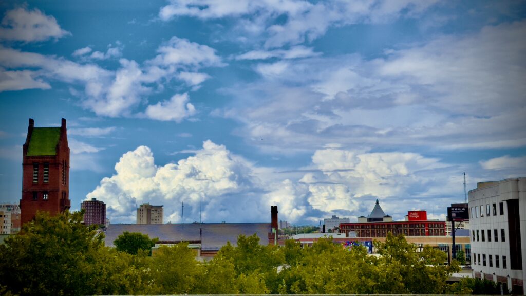 The image captures a cityscape view of Baltimore from an elevated vantage point, likely through a window. The foreground features lush green trees, while the middle ground includes a mix of historic and modern buildings. A tall, red brick tower with a green roof stands prominently on the left side of the image, contrasted against a backdrop of towering cumulus clouds. The sky is a vibrant blue, filled with a mix of fluffy white clouds that appear to be rolling in, possibly signaling an approaching storm. The scene exudes a sense of calm and beauty, with the natural elements juxtaposed against the urban environment.