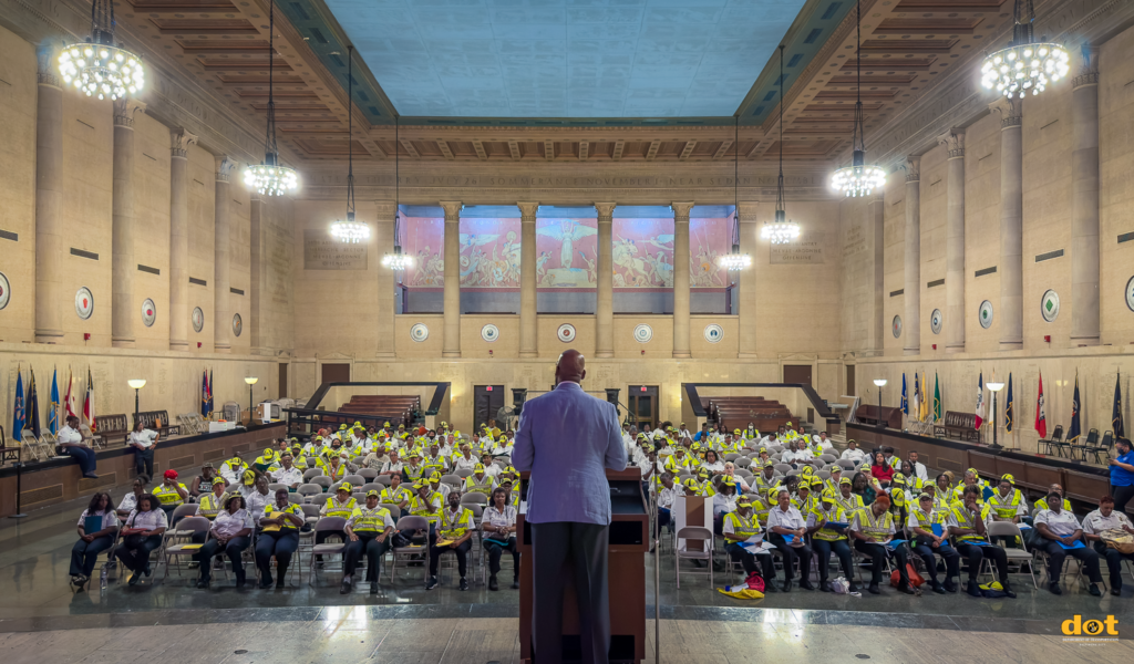 The image captures a large indoor assembly at the annual Crossing Guards Symposium, viewed from behind the speaker, Alan Robinson, as he sets the tone for the day with his energetic presence. The event takes place in a grand hall, featuring tall, ornate columns along the walls, adorned with circular emblems and flags between them. The ceiling is decorated with elaborate chandeliers that cast a warm glow over the room. The audience, mostly crossing guards wearing bright yellow vests and caps, is seated in rows of chairs, facing the speaker. Some attendees are taking notes or holding materials, fully engaged in the proceedings. At the far end of the hall, large murals or panels add a historical and artistic backdrop to the formal setting. The Department of Transportation (DOT) logo is visible in the bottom right corner, indicating the event's focus on transportation and safety.