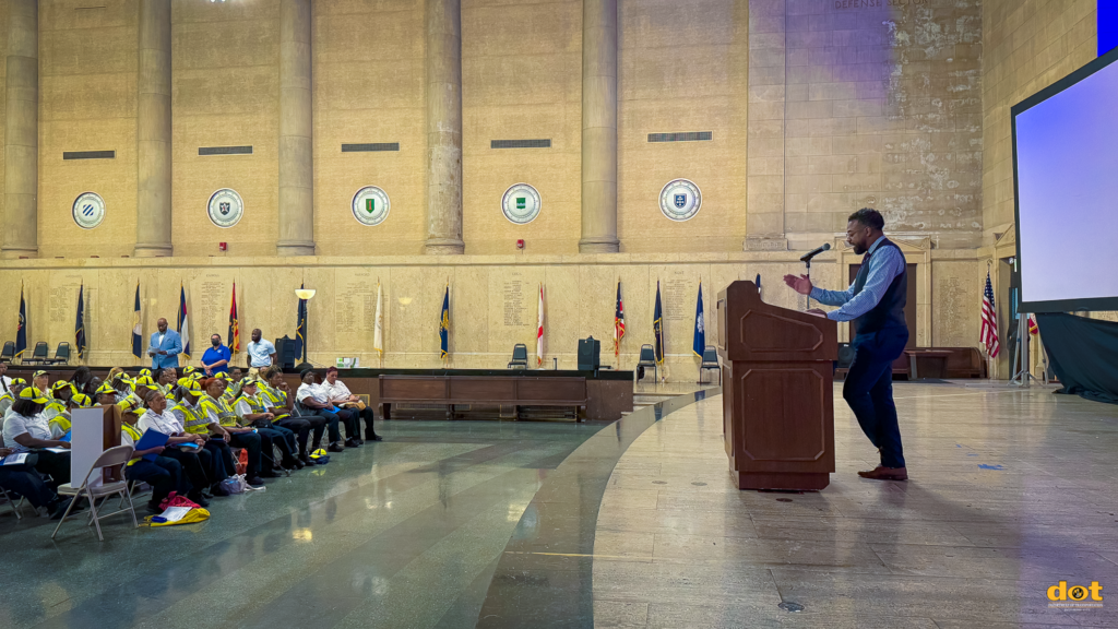 This image shows Tavon Braxton speaking at the podium during the annual Crossing Guards Symposium. He is dressed in a dark vest and light blue shirt, addressing an audience of crossing guards, many of whom are wearing bright yellow vests and caps. The setting is a grand hall with tall columns, circular emblems on the walls, and various flags displayed behind the audience. The audience appears attentive, and the overall atmosphere is formal and focused, reflecting the importance of the event.