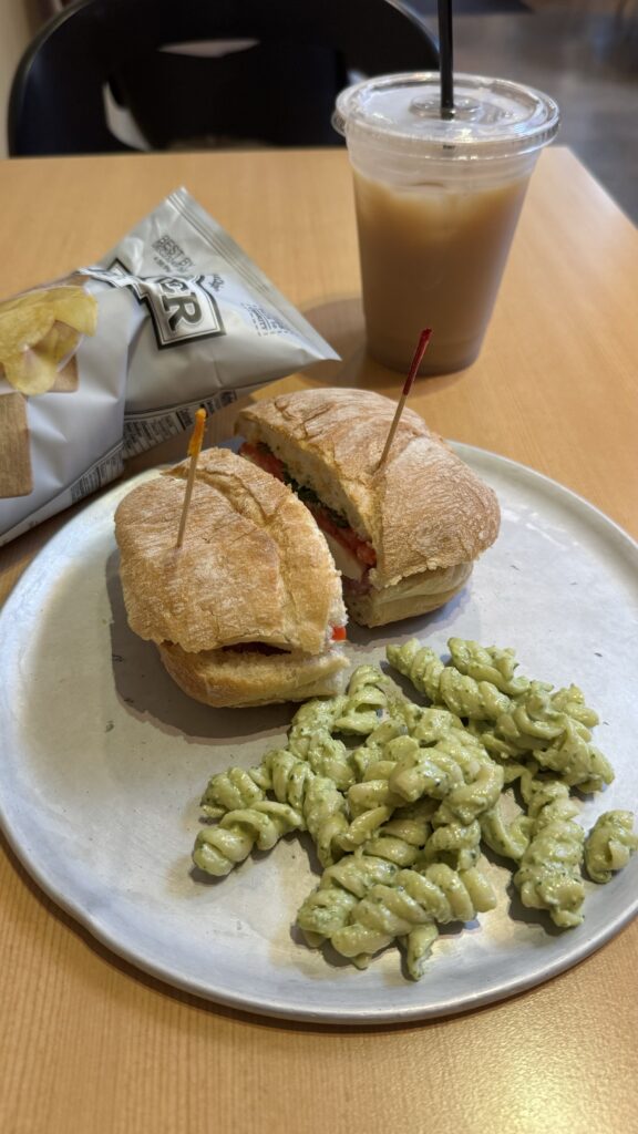 The image shows a meal set on a light wooden table, consisting of a sandwich, a side of pasta salad, a bag of potato chips, and a cup of iced chai. The sandwich, made by Rosina Gourmet, is served on a round plate and is cut into two halves, each secured with a toothpick. It appears to be a ciabatta roll with ingredients like tomato, greens, and possibly cheese and deli meat. To the side of the sandwich is a small portion of rotini pasta salad coated in a green pesto sauce. A bag of potato chips is partially visible next to the plate, and a clear plastic cup of iced chai with a black straw is placed behind the plate. The setting suggests a casual lunch, possibly at a café or sandwich shop.