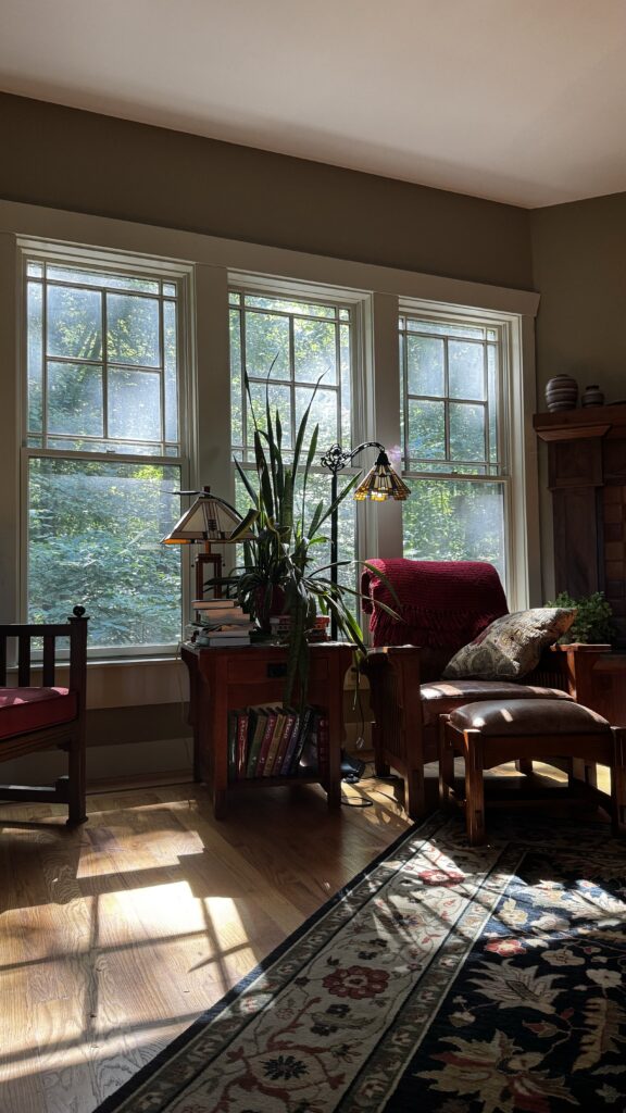 The image shows a cozy, sunlit corner of a living room. Sunlight streams through three large windows, casting warm light onto the hardwood floor and illuminating the room. A comfortable wooden armchair with a red cushion and a floral-patterned pillow is positioned near the windows, with a matching footstool in front of it. Next to the chair is a wooden side table holding a tall, leafy houseplant and a Tiffany-style lamp with a stained-glass shade. Books are stacked on the table and neatly arranged on the lower shelf. The room has a warm, inviting atmosphere, with natural light enhancing the earthy tones of the wooden furniture and the decorative area rug with a floral pattern that partially covers the floor. The overall scene evokes a sense of calm and comfort, ideal for reading or relaxing.
