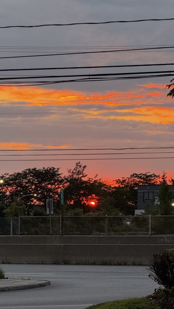 The image captures a vibrant sunset as seen from the Holiday Inn in Montreal. The sun is low on the horizon, glowing a deep red-orange as it sets behind a line of trees. The sky is a mix of warm oranges and reds with patches of soft gray clouds. Power lines stretch horizontally across the upper part of the image, adding an urban element to the scene. Below, a road and some greenery are visible, partially lit by the remaining daylight. The overall scene blends the beauty of nature with the everyday elements of the city.
