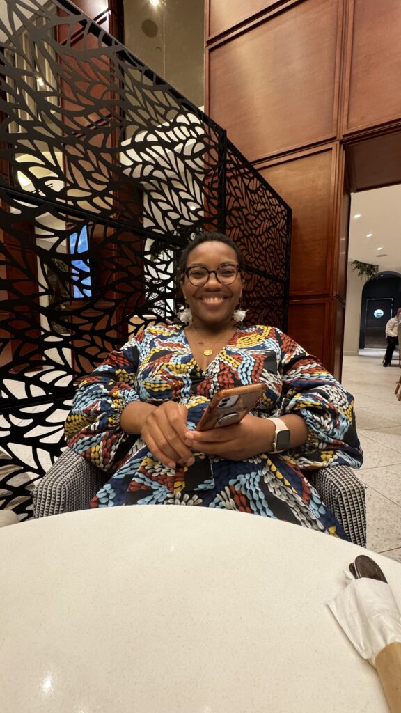  The image shows a woman, identified as Marilyne, smiling warmly while seated at a table during a dinner date. She is wearing glasses and a colorful, patterned dress with shades of blue, red, yellow, and white. Marilyne is holding a smartphone in her hands, resting them on the table. Behind her is a decorative black screen with intricate cut-out designs, which contrasts with the warm wood paneling of the restaurant’s interior. The atmosphere appears cozy and intimate, highlighting the special moment between her and the person she’s with on this dinner date.
