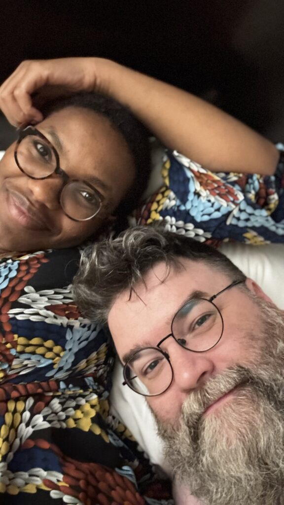 The image is a close-up selfie of a couple, Marilyne and Benjamin Jancewicz, lying together and smiling at the camera. Marilyne is on the left, with her head resting on her hand, wearing glasses and a colorful, patterned outfit in shades of blue, red, yellow, and white. Benjamin is on the right, also wearing glasses, with a full beard and a content expression. The photo captures a moment of closeness and warmth between the two, reflecting their connection and affection for each other.