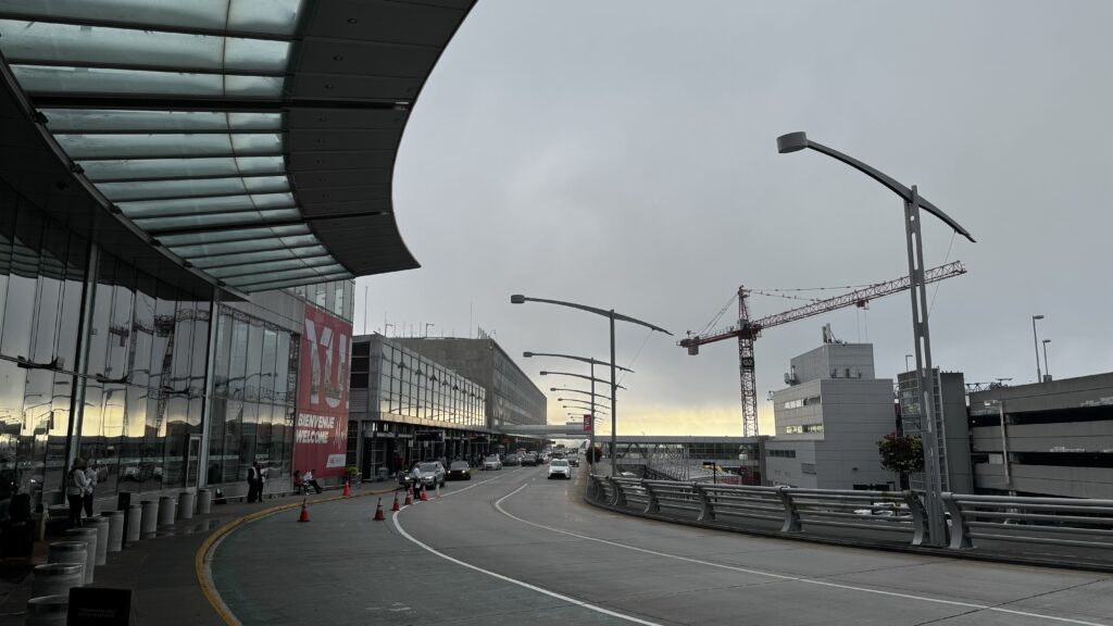 The image shows an exterior view of Montreal's airport terminal at dawn or dusk, with the sky overcast and the horizon glowing faintly with a hint of sunrise or sunset. The terminal building on the left features large glass windows reflecting the scene, with a curved glass canopy above the sidewalk. A prominent red and white sign on the building reads "Bienvenue / Welcome" in both French and English. Several vehicles are parked or moving along the curved road leading to the terminal entrance, and construction cranes are visible in the background, indicating ongoing development. The scene is calm, with a few people visible near the entrance, emphasizing the typical airport atmosphere.