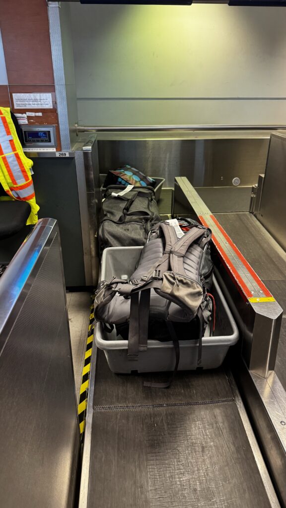 The image shows a set of backpacks and luggage bins being processed through an airport's baggage handling system. Two large backpacks are placed inside gray plastic bins on a conveyor belt, ready for checking. One bag is black, and the other is gray with visible straps and padding. A partially visible airport worker, identifiable by a neon yellow safety vest with reflective orange stripes, stands to the left. The setting is industrial, with metal surfaces and yellow and black safety tape lining the edge of the conveyor belt, creating a typical airport baggage handling scene.