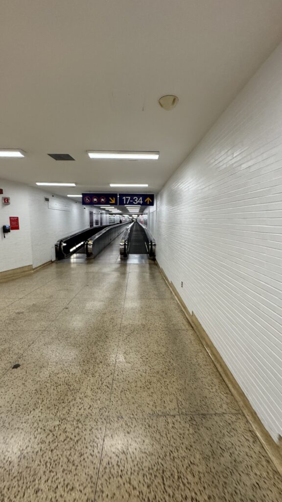  The image shows a long, empty airport corridor leading to gates 17-34, with two parallel moving walkways running down the center of the hall. The walls are white with a tiled texture, and the floor is speckled in shades of brown. Overhead, a sign displays gate numbers and symbols indicating accessible routes, no smoking, and no mobile phones. The walkways are lit by ceiling lights, creating a bright but slightly sterile atmosphere. The corridor appears to stretch far into the distance, with no visible people or activity, emphasizing the emptiness and quiet of the space.