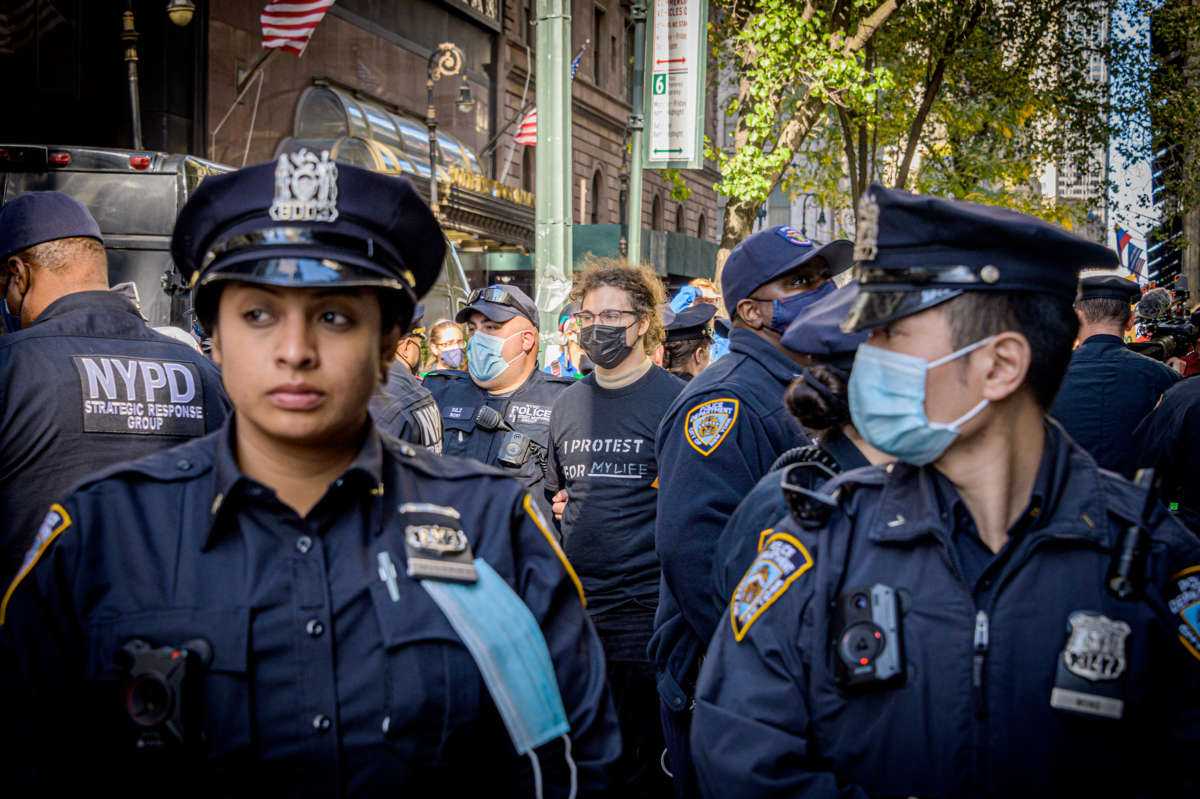 A group of NYPD officers, some wearing masks, stand in a crowded city street during a protest. In the background, a protester wearing a black shirt with the words "I PROTEST FOR MY LIFE" is seen amid the officers. American flags and city signs are visible, adding to the urban setting. The scene reflects tension, with police maintaining a strong presence.