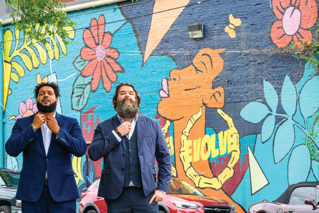 Two men in navy suits, standing confidently and adjusting their collars, pose in front of a vibrant mural featuring a large, colorful portrait of a woman and flowers. The word "Evolve" is visible on the mural behind them.