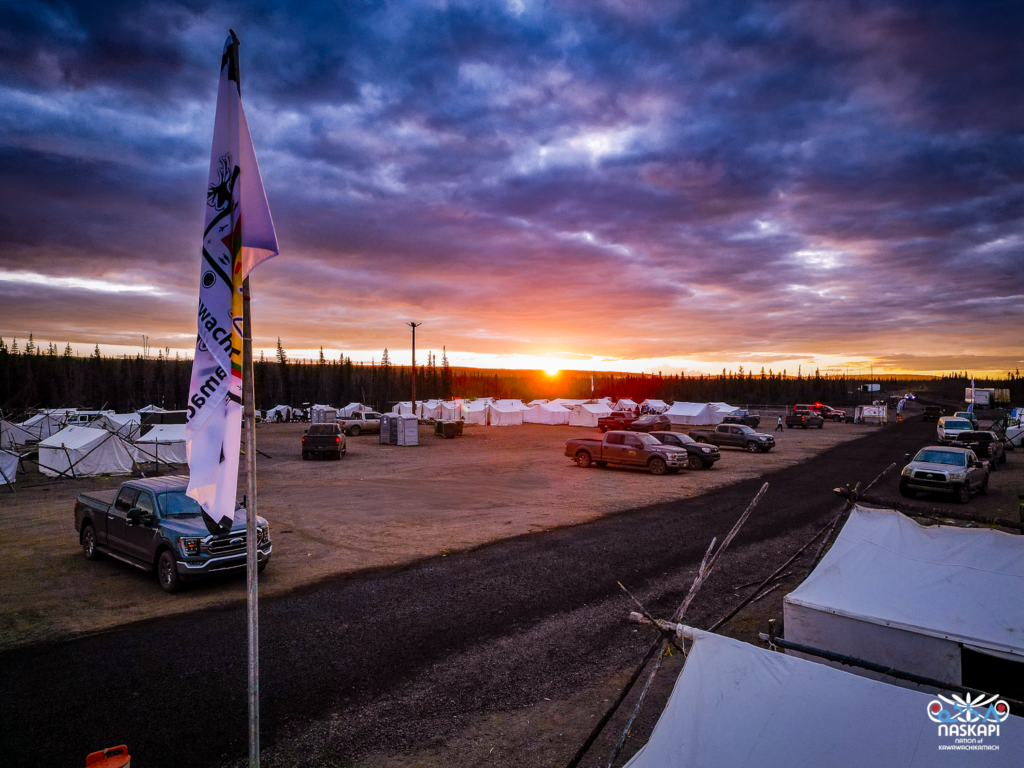 A wide-angle shot of a vibrant sunset over the tent village. In the foreground, the Naskapi flag flutters in the breeze, and a row of trucks is parked. The sky is awash with purples, oranges, and reds as the sun dips below the horizon, casting a warm glow over the rows of white traditional tents.