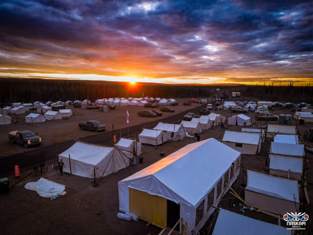 Another wide-angle drone shot of the tent village at sunset. The sky is filled with dramatic, colorful clouds, and the village below shows a peaceful scene with tents organized into neat rows. Trucks are scattered throughout the area, and the horizon stretches endlessly into the darkening forest.