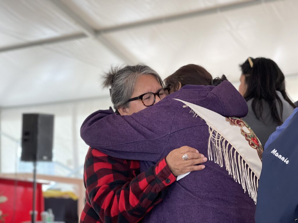 A tender moment where two women share an emotional hug during the session. One wears a purple jacket with a white fringed shawl, and the other is in a red and black checkered jacket. Both are visibly moved by the session.
