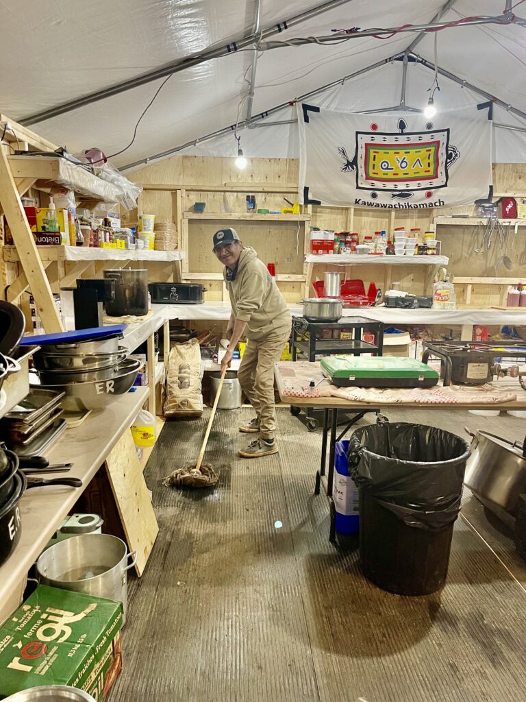 A man mops the floor in a large kitchen area set up in a tent. The space is filled with shelves of supplies, pots, and cooking equipment, with a banner in the background that reads "Kawawachikamach." He is smiling as he works, surrounded by the organized yet busy setting.