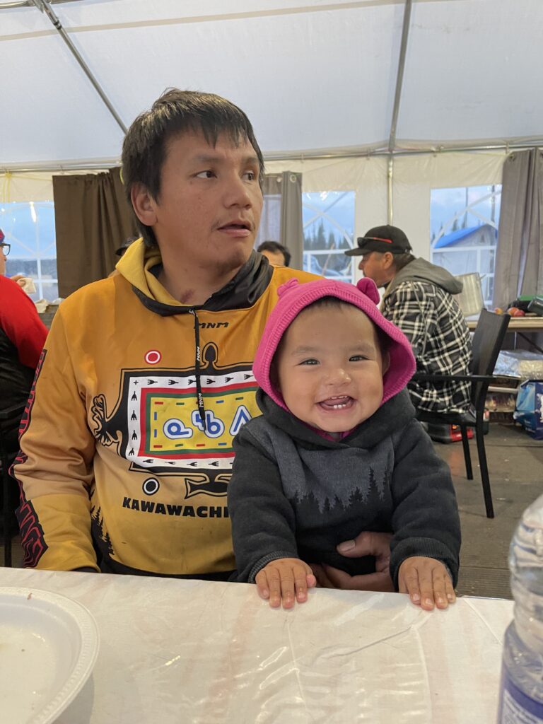 This image shows a man sitting at a table with a baby in front of him. The man is wearing a yellow hoodie with colorful designs, including text that reads "Kawawachikamach." The baby, wearing a dark hoodie with a pink hood, is smiling brightly, showing small teeth, and appears to be in a cheerful mood. They are inside a tented area, likely during a communal meal or gathering, with other people seated in the background. The scene radiates warmth and happiness.