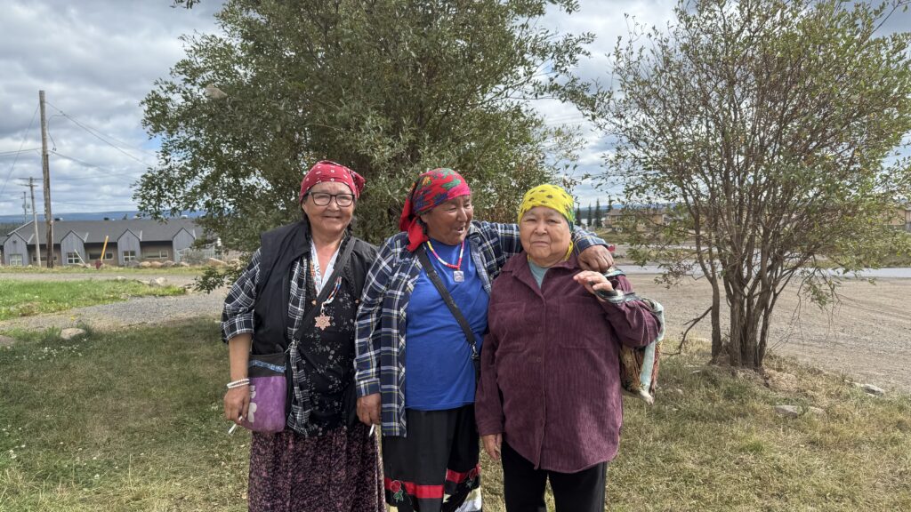 Naskapi Elders: Three Naskapi women standing together outside. All three are wearing colorful headscarves – one red, one multicolored, and one yellow. They are smiling and standing closely together, one with her arm around the other two. Behind them, there are some trees and small buildings under a cloudy sky.