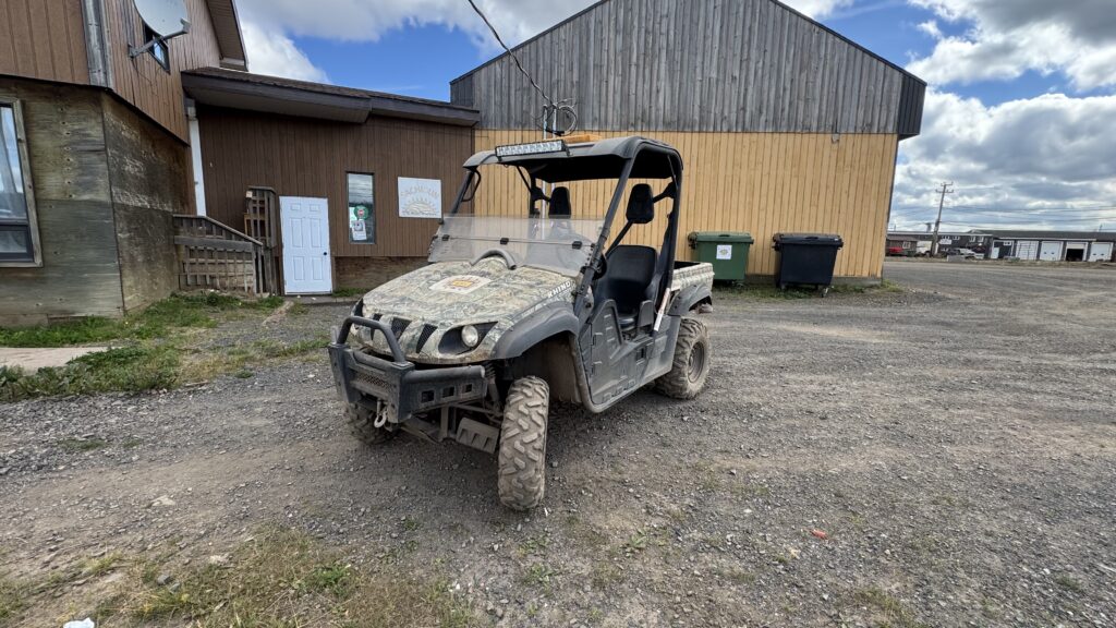 A rugged side-by-side vehicle parked on a gravel lot in front of a building with brown wood siding. The vehicle, a Yamaha Rhino with a camouflage paint job, has visible dirt on its tires and body. There is a white door on the side of the building in the background, along with a few windows and a satellite dish. A couple of green trash bins are also visible near the building. The sky is partly cloudy with patches of blue.