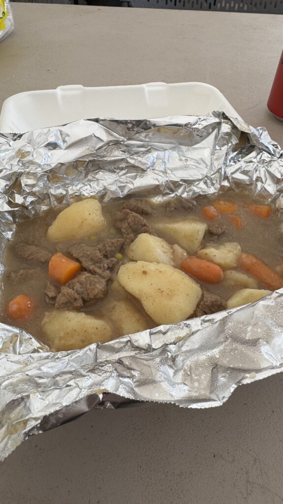 A close-up shot of a serving of caribou stew inside a takeout container lined with aluminum foil. The stew contains large chunks of tender caribou meat, potatoes, carrots, and peas, all immersed in a savory broth. The rustic presentation showcases a hearty, traditional dish, highlighting its rich, homemade quality. The background features a plain, light-colored table surface, with part of a red soda can visible on the right side of the image.