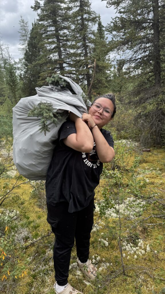 A woman, smiling, carries a large bag of spruce boughs on her shoulder through a forest. She wears glasses and black clothing, with the surrounding landscape full of lush green moss and trees.