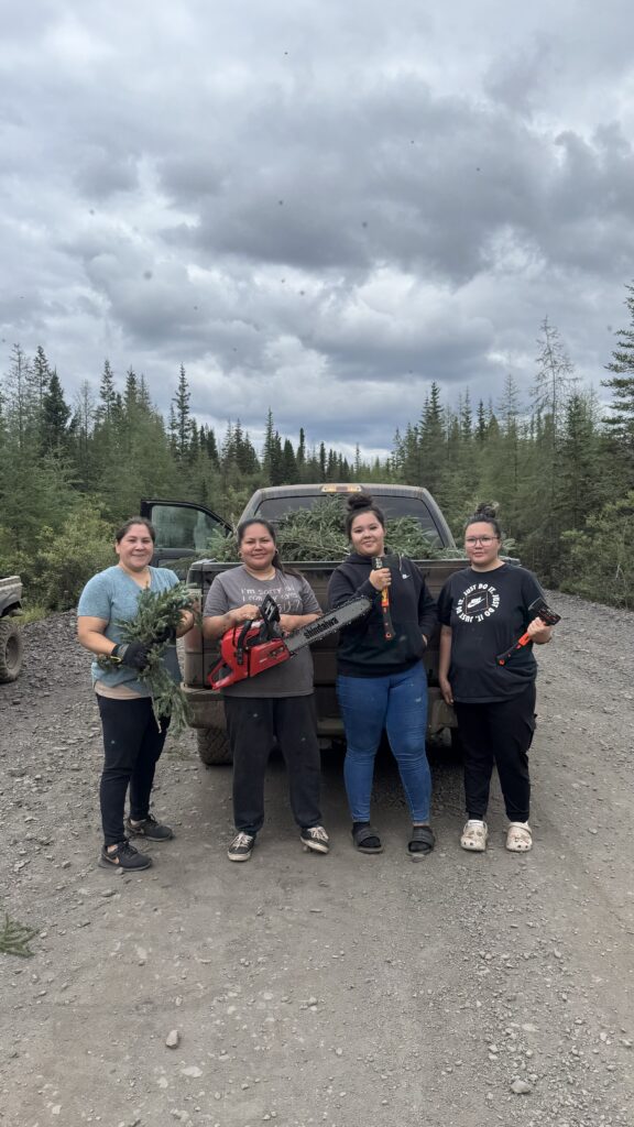 Four women standing on a gravel road, holding various tools, including chainsaws and axes. They pose proudly in front of a truck filled with spruce boughs, with a cloudy sky and forest in the background.