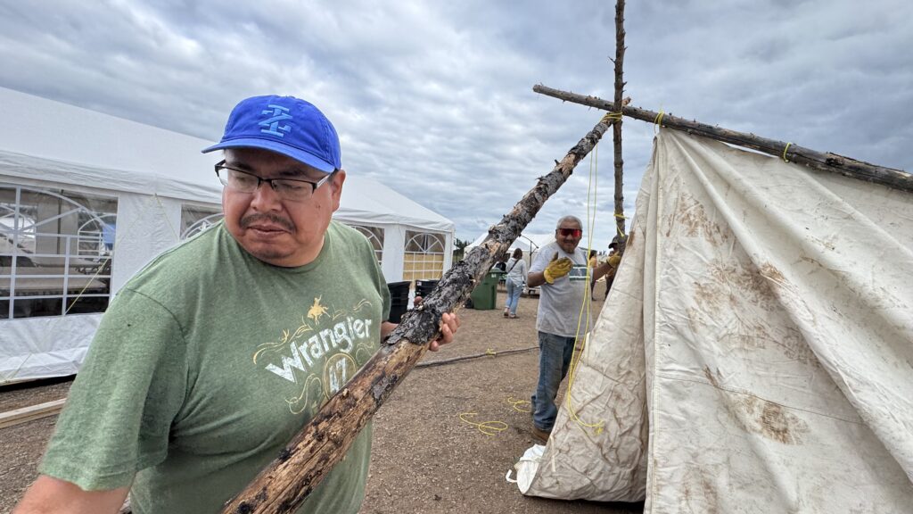 A man wearing a green "Wrangler" T-shirt and blue baseball cap is seen carrying a sturdy wooden pole as part of tent setup. In the background, another man wearing protective sunglasses and gloves is assisting with the assembly of a canvas tent supported by wooden poles, while other tents and a white pavilion stand under a cloudy sky.