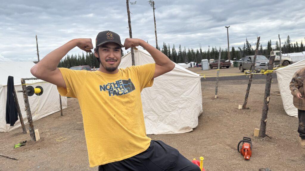 A young Naskapi man is playfully flexing his biceps and smiling for the camera. He is wearing a bright yellow "The North Face" T-shirt and a black baseball cap with a New York Yankees logo. Behind him, a campsite is in progress with white tents arranged in rows.