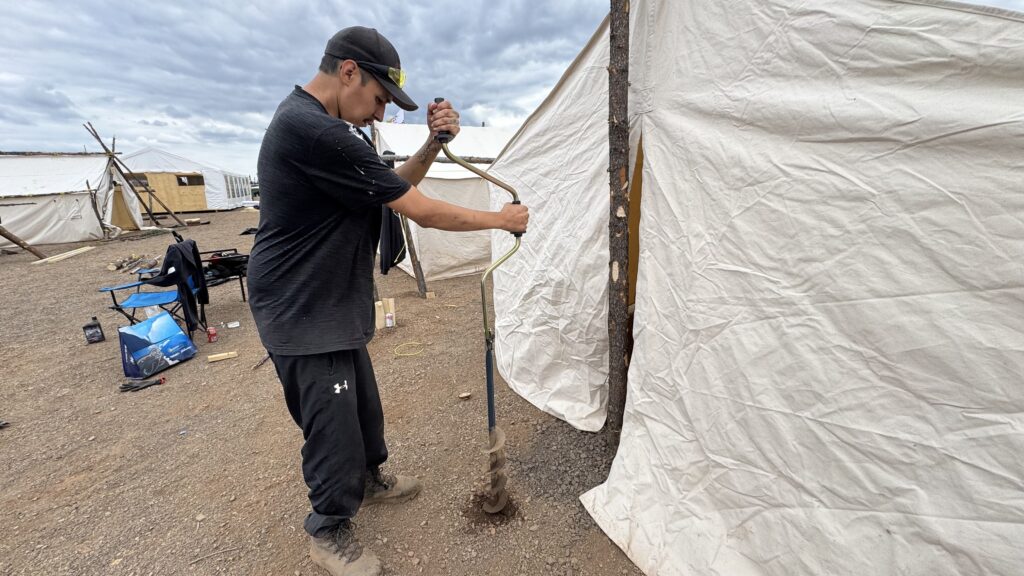 A Naskapi man, dressed in a black Under Armour T-shirt, is seen using an auger to drill holes in the ground next to a traditional canvas tent. The man concentrates on the task as he works to secure the tent structure. Behind him, various tools and tents are scattered across the camp.