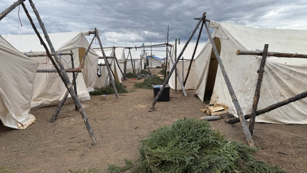 A row of traditional tents set up by the Naskapi, framed by wooden poles tied together with rope. The ground is covered in spruce boughs, and the tents are arranged in a linear formation, creating a clear path between them. The sky is overcast, giving the scene a muted, natural tone. The tents, made of off-white canvas, are part of a camp set up for the elders.