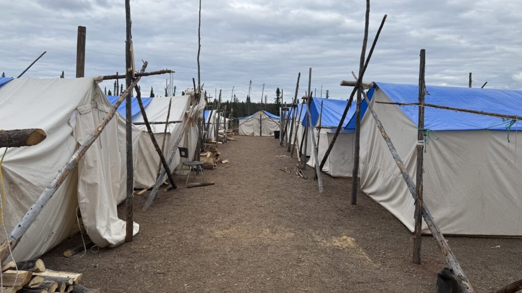 Another view of the camp, showing more traditional tents lined up in rows. The tents have wooden frames and white canvas walls, some covered by blue tarps for additional protection. The tents create a neat pathway in the middle, with logs and supplies scattered around the camp. The cloudy sky adds to the rustic feel of the camp setup.