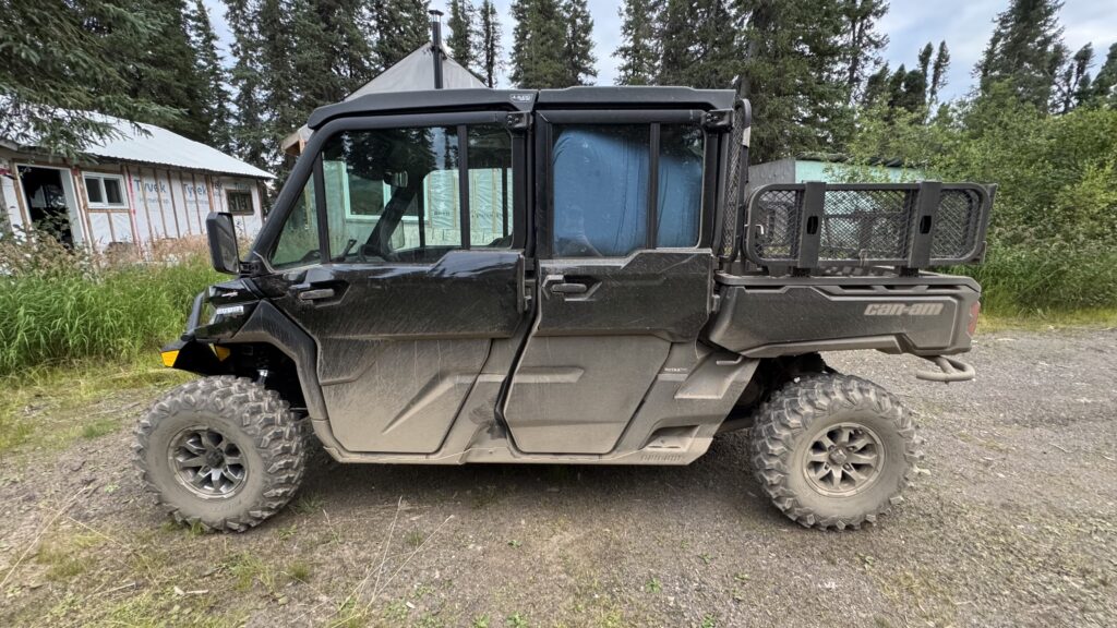 A side view of a large black Can-Am side-by-side utility vehicle parked on a dirt area surrounded by tall grass and trees. The vehicle is muddy from off-road use and features large rugged tires and a cargo bed in the back. The background includes a partially constructed building with Tyvek wrap and metal roofing, indicating an outdoor rural setting.