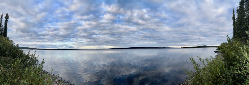 A panoramic view of Kachikayach Lake under a partly cloudy sky. The water is still, reflecting the clouds above, and the distant shoreline is visible, creating a tranquil scene. On both sides of the frame, green bushes and trees line the edges of the lake, while a small section of rocky shoreline is visible in the foreground. The horizon stretches across the middle of the image, with rolling hills in the distance.