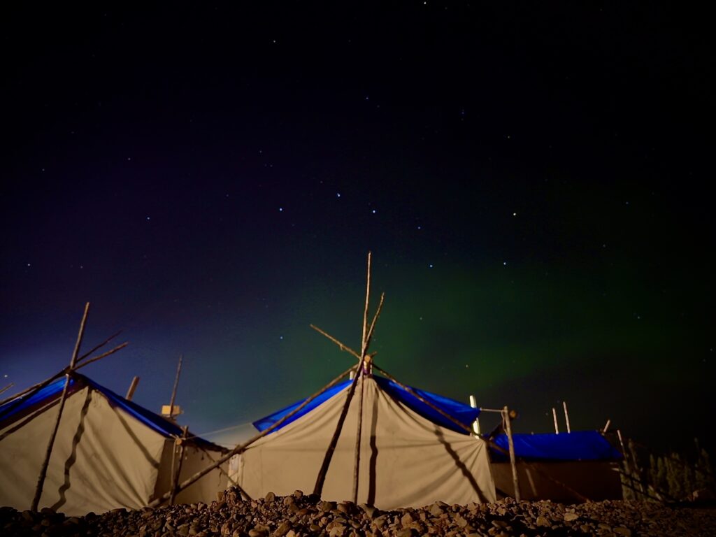 The greenish glow of the Northern Lights can be seen in the starry night sky above Naskapi tents, framed by tall, rustic wooden poles supporting the tents' structures. The tents have blue tops, adding a pop of color to the nighttime landscape.