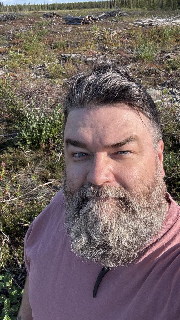 A close-up selfie of Benjamin Jancewicz with a gray beard, wearing a light pink shirt. The background reveals a rugged landscape of wild plants, tree stumps, and logs, indicating a natural or forested area. The man has a calm expression and the sun is shining, casting a soft light across his face.