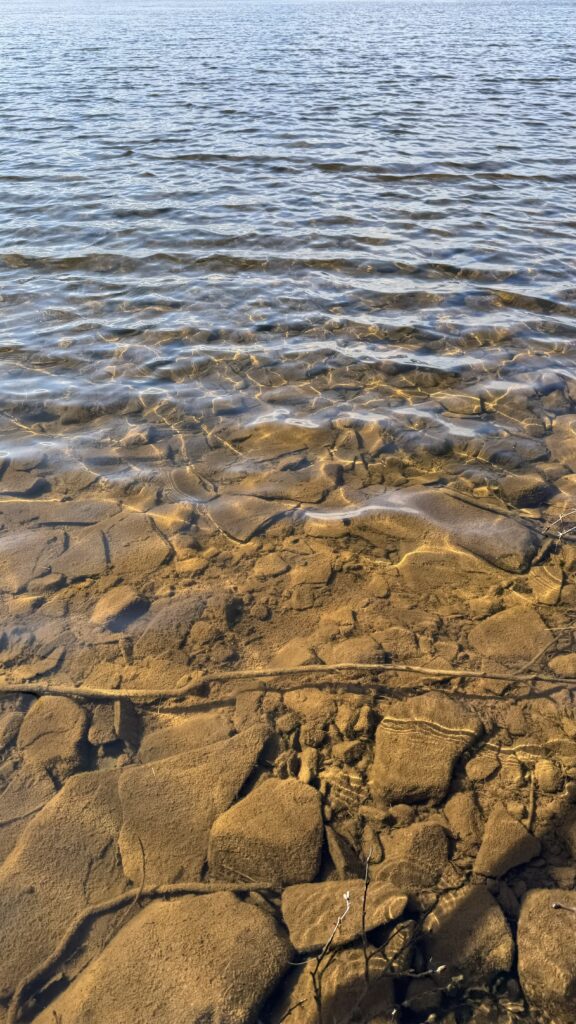 A shallow, clear lake with sunlight illuminating the rocks and pebbles at the bottom. The water ripples gently, creating a textured surface while maintaining visibility of the submerged, sandy-colored stones below.