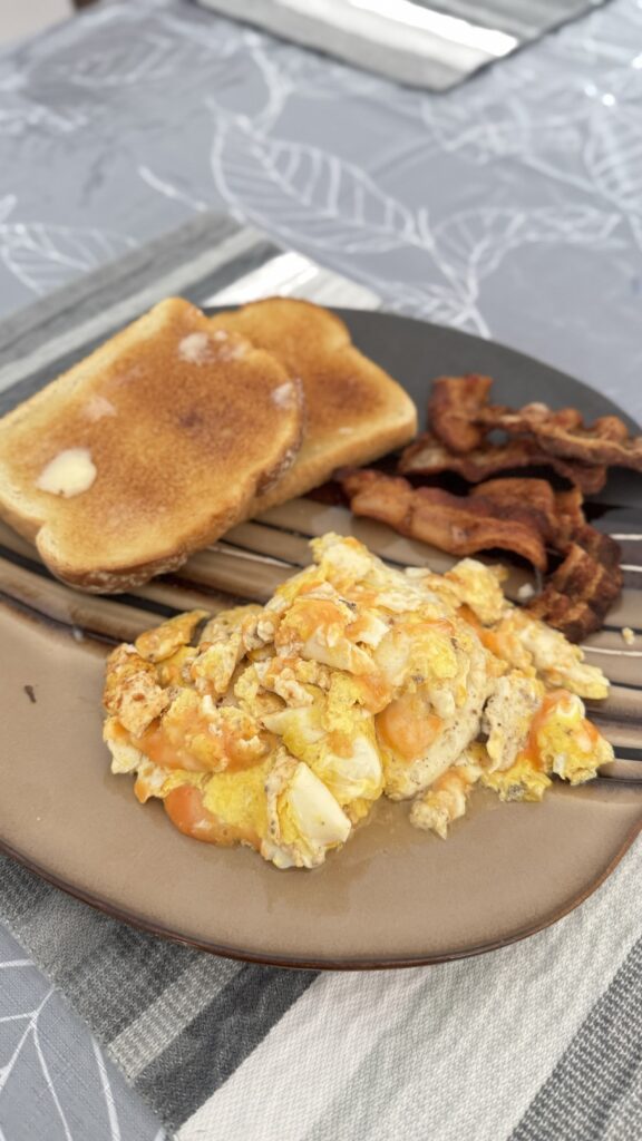 A plate of homemade breakfast consisting of scrambled eggs with melted cheese, crispy bacon, and two slices of buttered toast. The meal is set on a beige plate, resting on a gray and white patterned tablecloth. This breakfast was prepared after a return trip to Kawawachikamach for a shower and some quiet time.