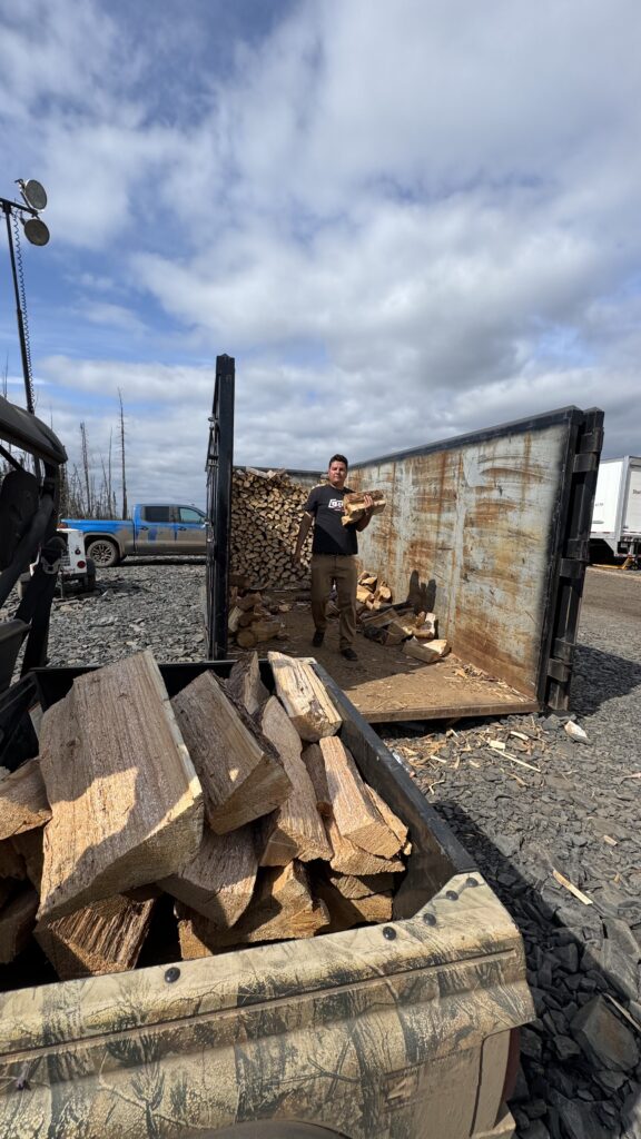 A man is standing in a large metal container filled with chopped firewood, holding a log in his hands. In the foreground, the back of a side-by-side vehicle is filled with more split firewood, ready for distribution. The scene is outdoors, under a partly cloudy sky, with another truck visible in the background. Benjamin Jancewicz is helping load and distribute firewood to various people at the camp.