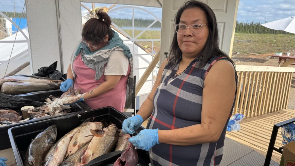 Image of two women cleaning fish and birds: Two women are focused on preparing food. One woman on the left is cleaning birds while wearing a pink shirt with a towel draped around her neck. The other woman is on the right wearing glasses, a plaid dress, and blue gloves as she works with a bird.