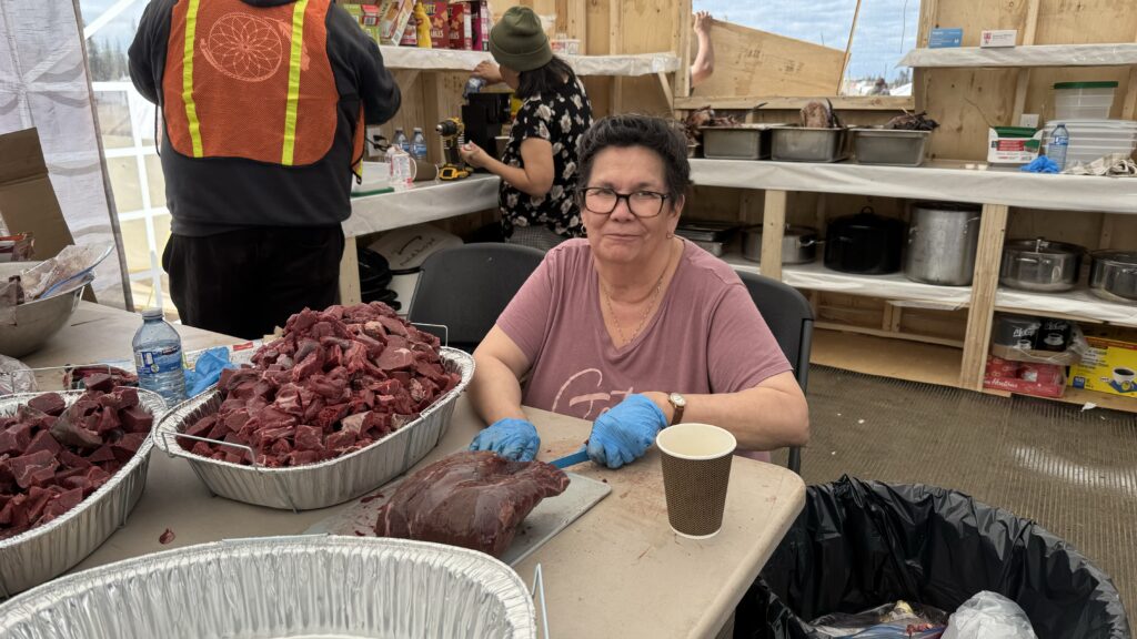 Image of an elder preparing meat in the Naskapi Kitchen: An elder sits at a table cutting a large piece of meat. In front of her are trays filled with chunks of caribou. She looks calmly into the camera, with kitchen supplies visible in the background.