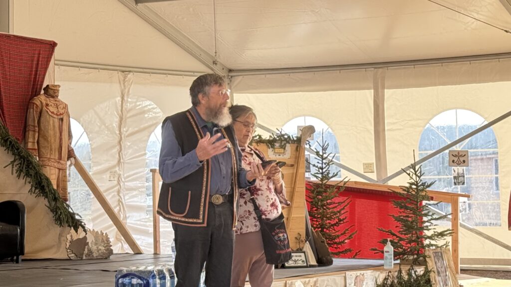 Benjamin Jancewicz's father, Bill Jancewicz, stands on a stage inside the big tent, leading a prayer alongside Maggie Mokoush-Swappie. They are dressed in traditional clothing, with Bill wearing a vest with colorful detailing, and Maggie holding a phone to read from. In the background, evergreen trees and traditional garments decorate the stage.