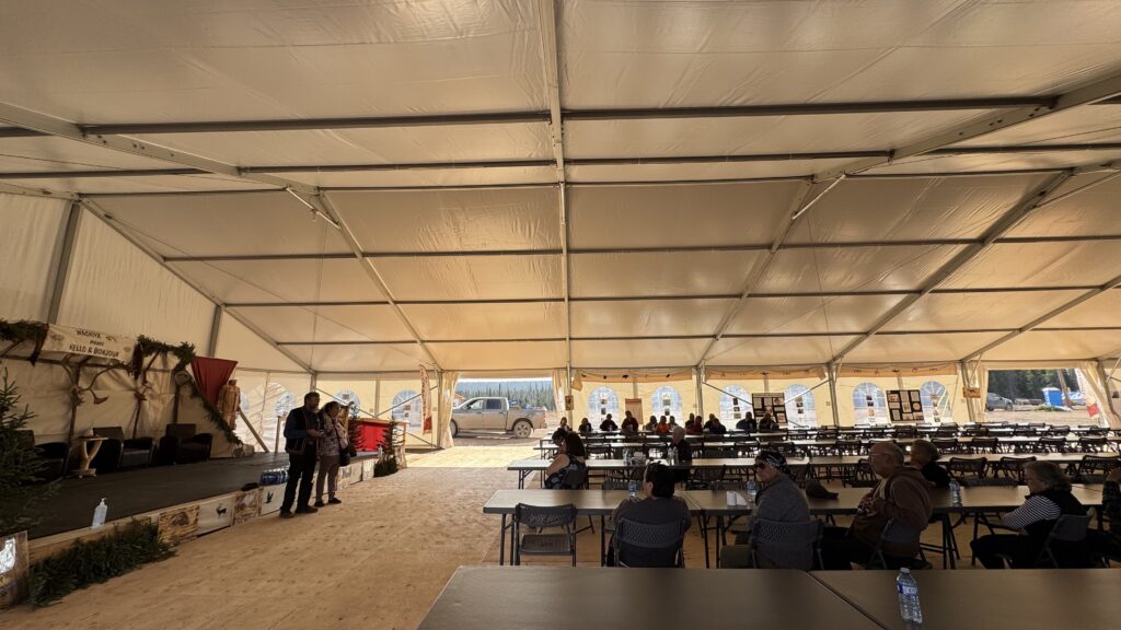 A second photo showing Bill Jancewicz and Maggie Mokoush-Swappie standing side by side on the stage. Bill reads from a phone while Maggie looks on thoughtfully. The soft sunlight filters through the tent, highlighting their traditional clothing and the peaceful atmosphere inside the big tent.