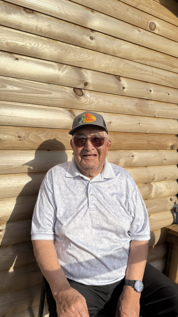 Image of an elder man in a gray shirt and a "Bass Pro Shops" hat: A close-up photo of a smiling elder man sitting outside a log cabin. He is wearing sunglasses, a gray polo shirt, and a watch. The warm sunlight casts soft shadows on the wooden wall behind him.