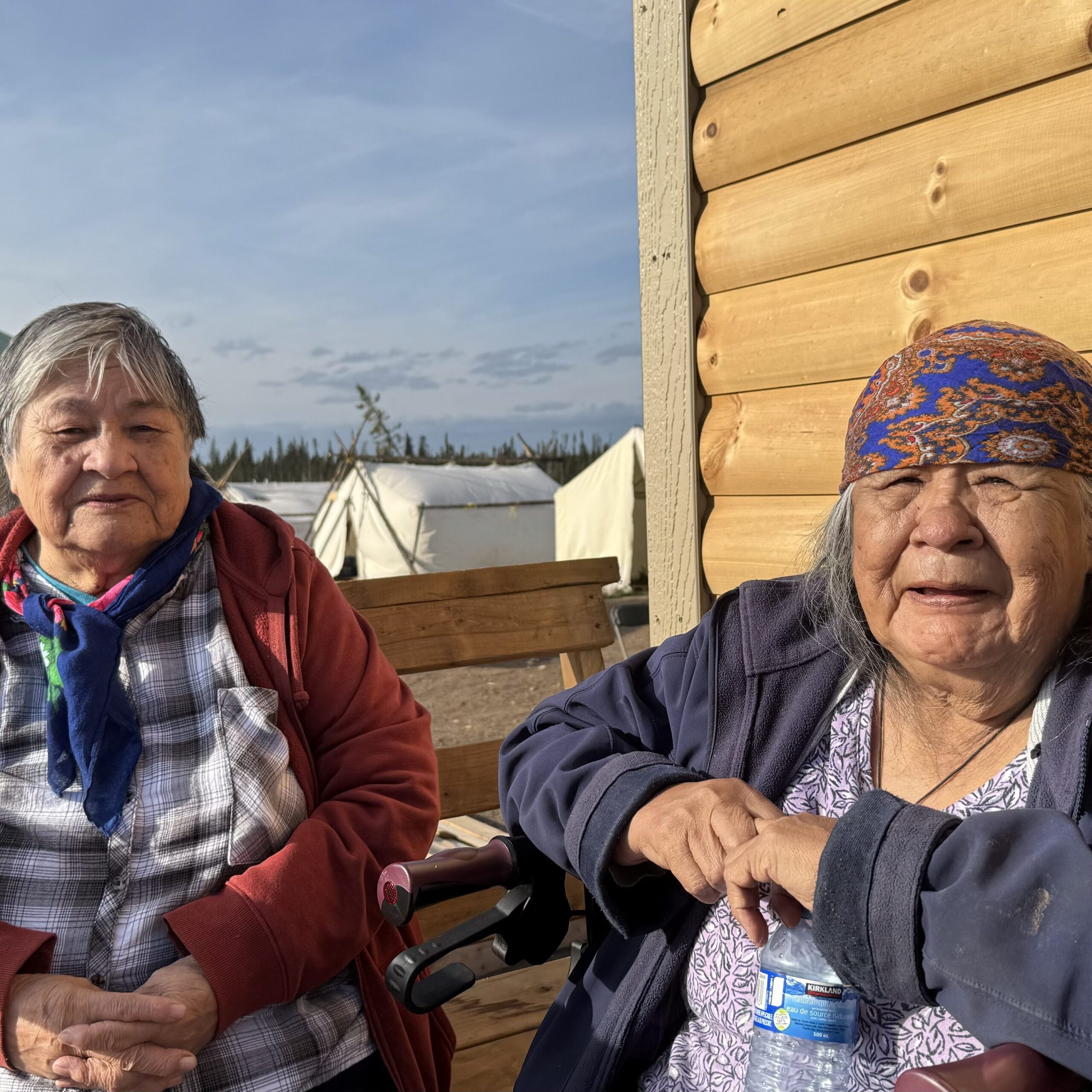 Two Naskapi elder women sitting together outside a log cabin: Two Naskapi elder women, one in a red jacket and the other in a navy-blue jacket and headscarf, sit together on a wooden bench outside a log cabin. They are bathed in golden sunlight, smiling softly as they enjoy the late afternoon.