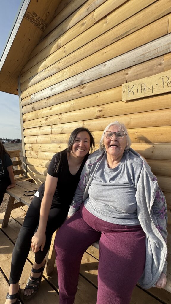 A young woman and an elder woman laughing together: A joyful moment captured as a young woman in a black shirt sits with an elderly woman who is laughing, wearing a gray sweater and purple pants. They sit on a bench outside a log cabin with the name "Kitty" engraved on a sign above.