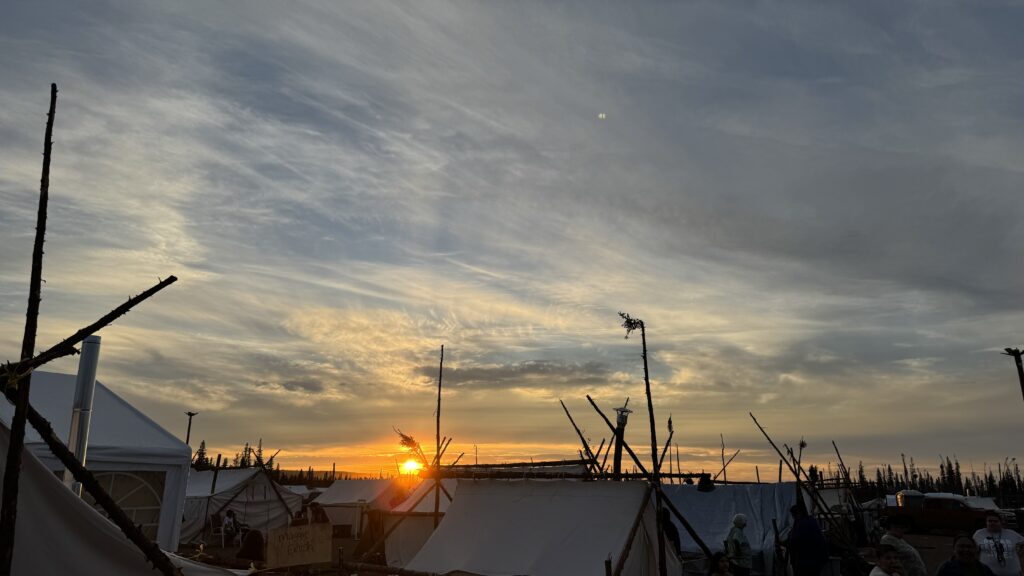 The sun sets behind a horizon of tents at the Naskapi camp, casting a warm golden light across the sky. The tents are framed by wooden poles, and a sign reading 'Magic English' is visible on one of them. The sky is filled with wispy clouds painted with soft hues of yellow, orange, and purple as the day transitions into evening. People gather around the tents, with some sitting and talking in the foreground, enjoying the peaceful atmosphere of the sunset.