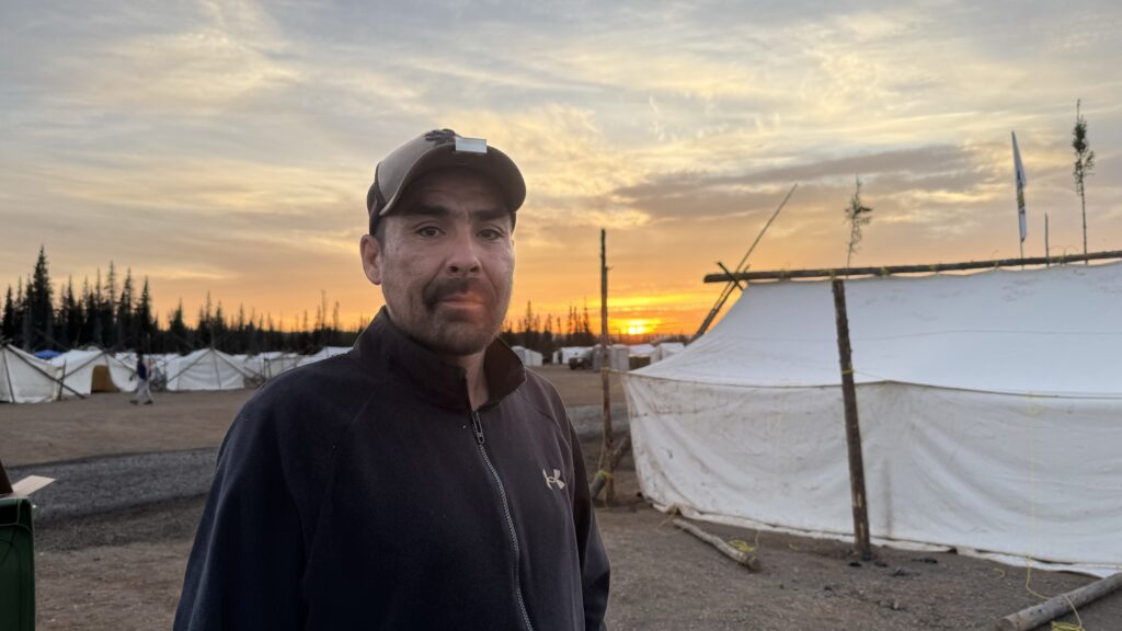 Natush Peastitute stands in front of a white tent at the Naskapi camp during sunset. He is wearing a black jacket and a baseball cap, looking directly at the camera with a calm expression. Behind him, the sun is setting, casting a warm golden glow across the sky, with soft clouds illuminated by the fading light. The camp's tents and poles are visible in the background, and the scene captures the peaceful yet industrious atmosphere of the camp where Natush has been working hard for over a week, building platforms and setting up beds for the elders.