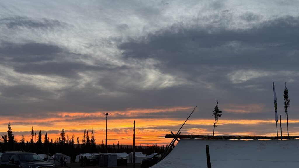 A colorful sunset over the camp with tents and wooden poles visible in silhouette. The sky is filled with vibrant shades of orange, pink, and purple as the sun sets behind the trees on the horizon. The camp is peaceful and quiet, with tents glowing softly in the fading light.