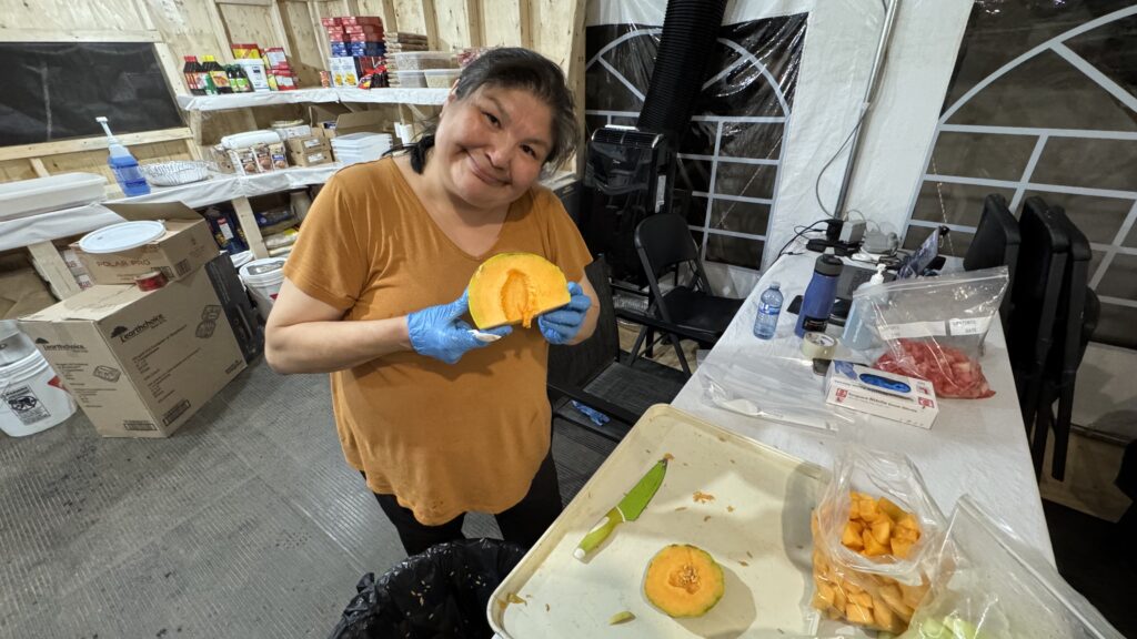 A close-up of a Naskapi woman in an orange shirt standing at a table with cut-up cantaloupe in hand. She holds half of the melon, showing its orange flesh while smiling warmly at the camera. She is wearing blue gloves and working in a kitchen space with food supplies in the background.