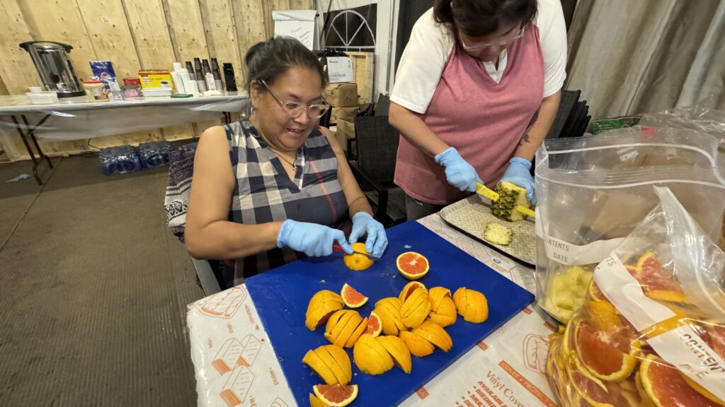 Two Naskapi women stand side by side, one cutting a pineapple and the other slicing oranges into small wedges. Both wear protective gloves and work on a large plastic board in the camp kitchen. The table is filled with fruits and bags of orange slices, with kitchen supplies stacked behind them.