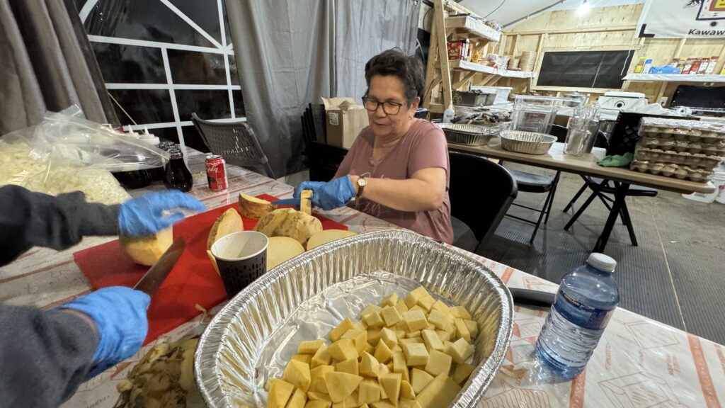 A Naskapi elder in a pink shirt sits at a table chopping vegetables with a large knife. The table is covered with foil pans full of cubed vegetables, and she appears focused on her work. The background shows the interior of the camp kitchen with stacked food supplies and kitchen equipment.