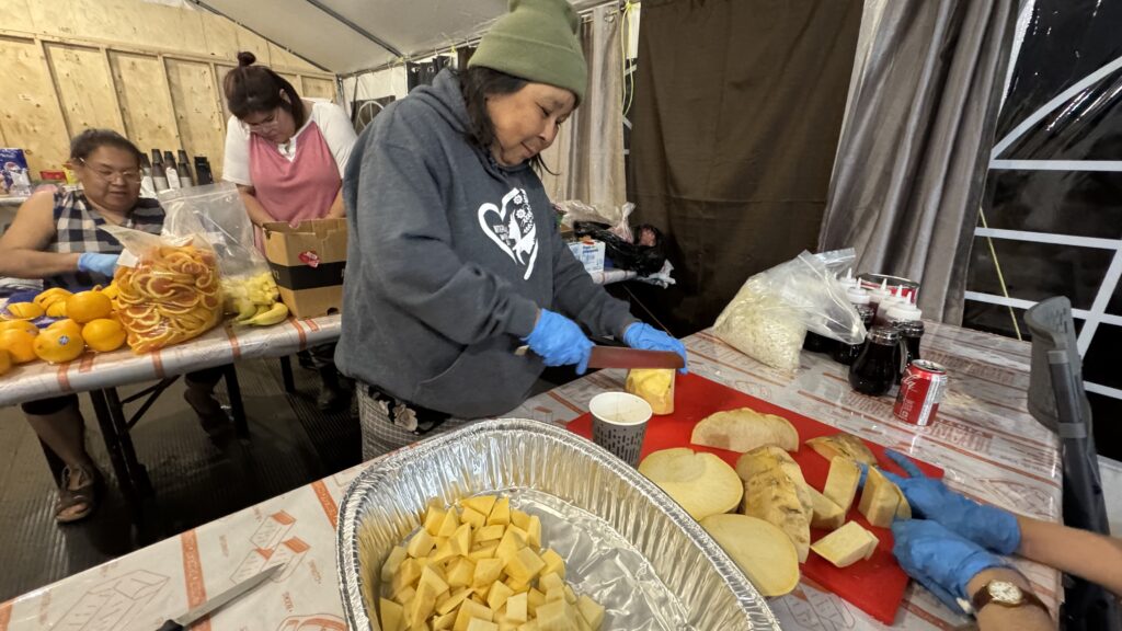 A group of Naskapi women in the kitchen, preparing food. Two of the women, one in an orange shirt and the other in a plaid top, smile while chopping various fruits and vegetables. They work amidst stacks of food supplies in the camp kitchen, surrounded by plastic containers, knives, and other kitchen tools.