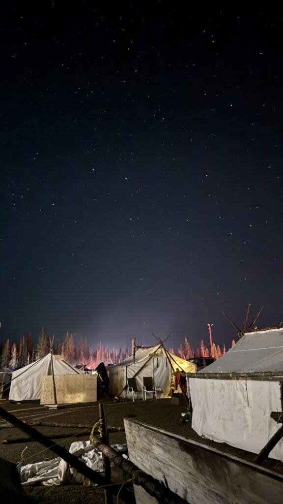 A breathtaking night sky filled with stars, with traditional tents below, illuminated softly. The campfire's glow in the background adds a warm light, while the sky remains clear and star-filled.